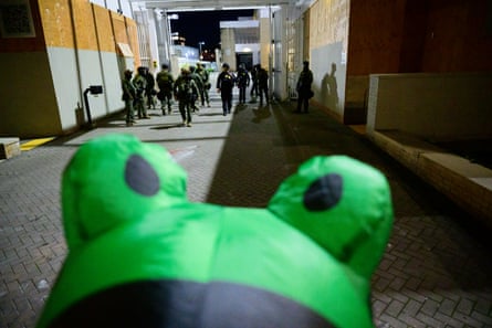 Frog costume in foreground with Ice agents beyond, down what appears to be an alley.