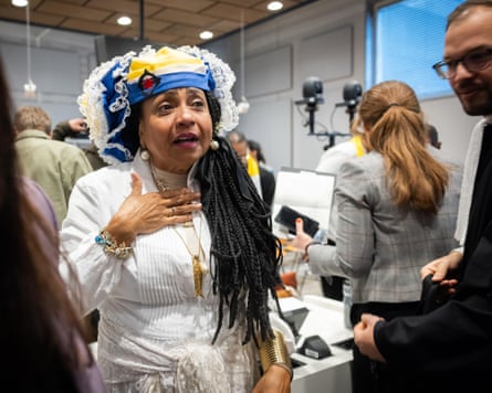 she holds one hand to her chest as she speaks. She has long braids and wears a white dress with lace trim, a gold necklace and bracelets, and a traditional-style yellow, black and white lace-trimmed headdress. People with cameras and microphones are circulating in the background.