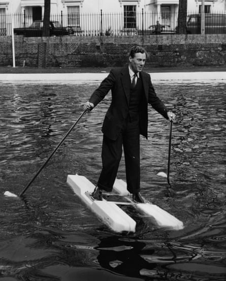 Black and white photograph of MW Hulton demonstrating his seashoes and duckfoot propellers on the Grand Union canal. He is in a dark suit and standing on what look like two wide skis, although they are joined by a small platform under his feet. The complete the ski look, he is holding what could be ski poles in his hands.
