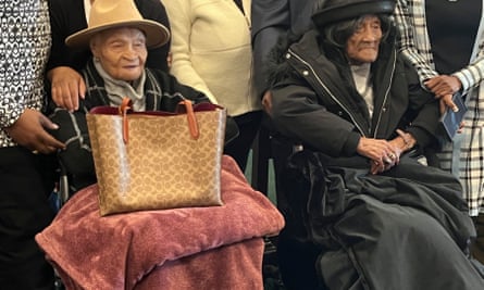 Two older Black women side by side in wheelchairs, backed by a number of people in business dress.