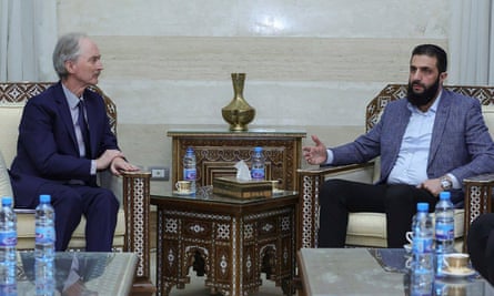 they sit formally in armchairs behind a carved wooden table. Sharaa wears an open jacket and white shirt.