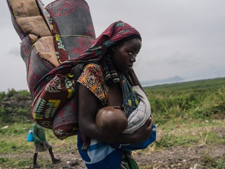 A woman in a grassy landscape carries a baby and large rolls of what appears to be bedding on her back