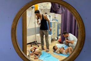two young children lie on mats on the floor in a hospital room; a man stands over one holding a drip and another man is kneeling by the other child; both men are in casual clothing and look like family members rather than medical personnel. Both children have blood on them and there is blood smeared across the floor. The image is framed through a circular window