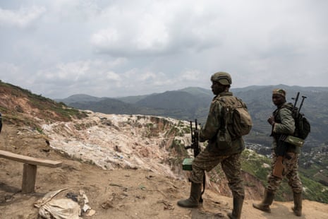 Two M23 soldiers in military gear stand at the edge of a Rubaya mining pit with mountains behind.