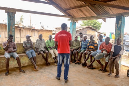 Issah Seidu stands with his back to the camera talking to a group of men sitting on benches under a shelter.