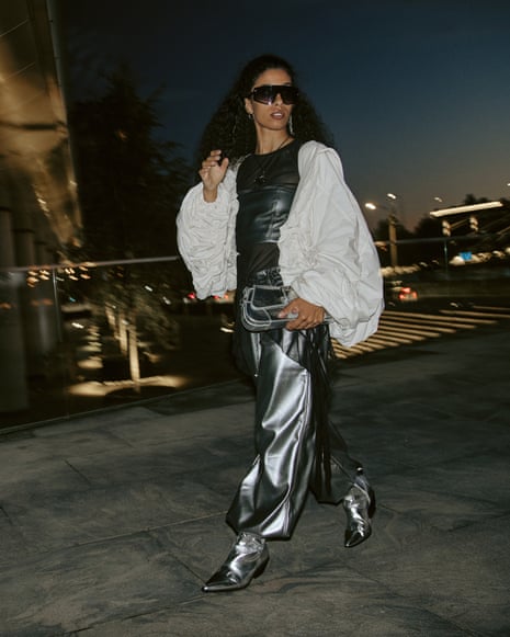 Woman walking in silver metallic pants and a wavy flowing white jacket with sunglasses at night on a concrete bridge or path