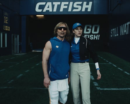 Powell in a muscle tank stands with a woman in a windproof jacket & baseball cap in a stadium