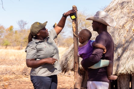 A woman holds a light on a post watched by a man holding a baby. They are outside in a savannah landscape next to a small thatched dwelling
