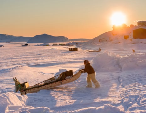 Man pulls loaded sledge across ice. Other sledges and scattered houses in background