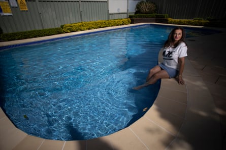 A woman in a white T-shirt and shorts, sitting at the edge of a back yard swimming pool with her legs dangling in the water.