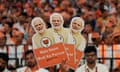 A hand holds three cardboard cutouts of Narendra Modi with party supporters out of focus in the background