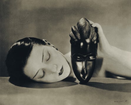 A white woman rests her right cheek on a tabletop while her left hand balances a black African face mask on the table