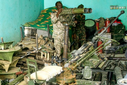 A man in camouflage uniform stands behind a gun in a room filled with military equipment.
