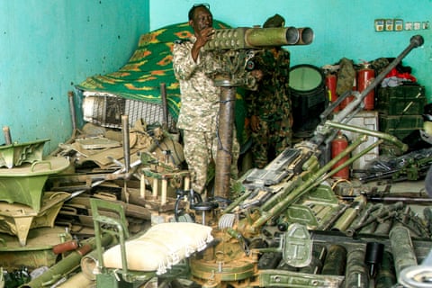 A man in camouflage uniform stands behind a gun in a room filled with military equipment. qhxidiqxkiqteinv
