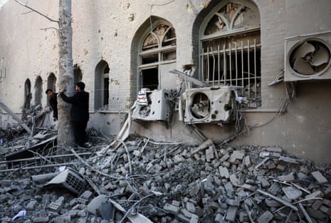 काले कपड़े पहने दो व्यक्ति एक क्षतिग्रस्त इमारत के पास एक नंगे सफेद पेड़ के बगल में मलबे की जमीन पर खड़े हैं।