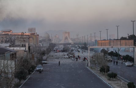 Broad landscape view of debris in the street from a destroyed building, on a road leading to Azadi Tower (an inverted Y-shaped structure) in the distance. Vehicles, some damaged, are stopped in the road as people and rescuers walk around