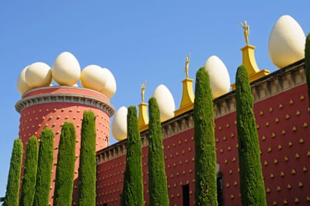 it is a pinkish-red building with huge white stone eggs and gold figurines along the roof, a circular tower, and tall thin green trees in front. The sky behind is bright blue.