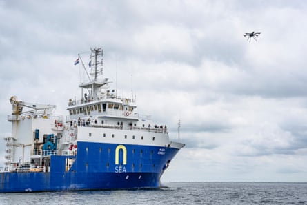 A large blue and white ship out at sea while a drone flies overhead
