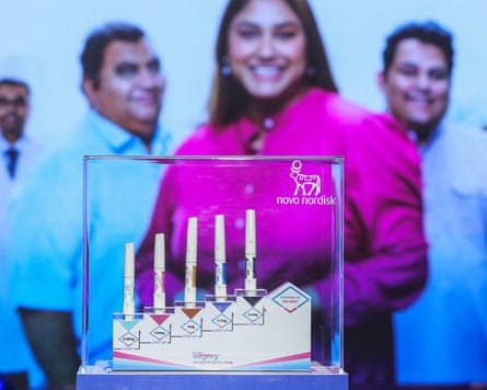 Wegovy injection pens are in a glass case in the foreground. Behind them is a poster of four people