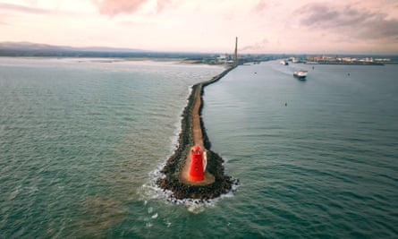A striking view of a red lighthouse at the very end of a narrow outcrop, contrasted with the blue-green sea.