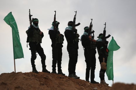Masked Hamas militants stand armed atop a dirt mound beside green flags in Gaza