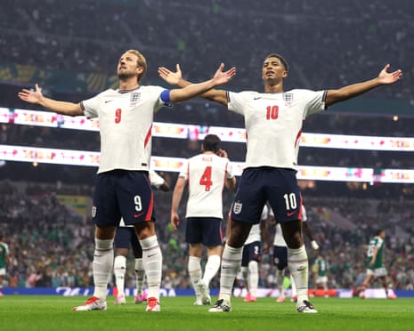 Jude Bellingham and Harry Kane in white England kits celebrate with arms outstretched on a pitch