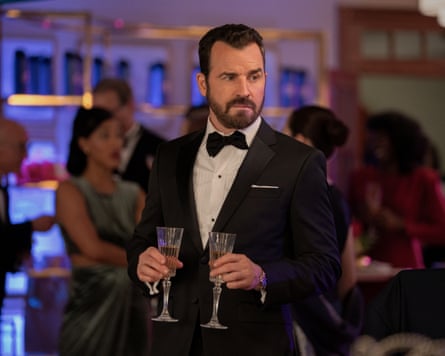 Justin Theroux in a dinner suit and bow tie, holding two champagne flutes, with guests blurred in the background.