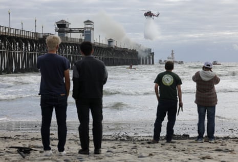 people stand on a beach by the ocean facing a pier with plumes of smoke rising from it