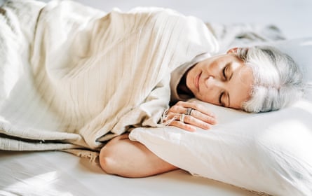 A middle-aged woman with short gray hair sleeps peacefully in bed, covered with a light blanket, her hand to her face, in a room bathed in bright morning sunlight.