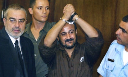 Marwan Barghouti holds his handcuffed arms in the air. A guard and two other people look on
