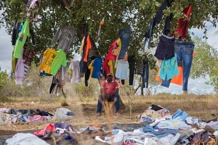 A man sits under a tree which is hung with items of clothing. More clothes are spread out on the ground around him