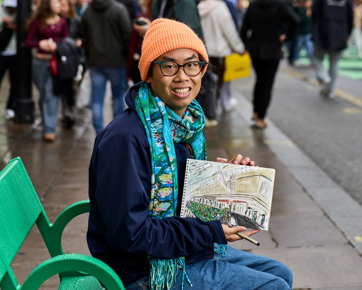 Daniel Te sits on a bright green bench holding out his sketch of the street. He wears a blue and green patterned scarf and orange woolly hat.