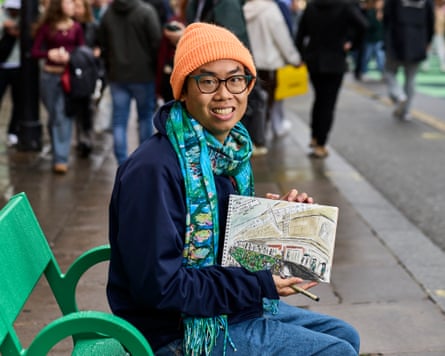 Daniel Te sits on a bright green bench holding out his sketch of the street. He wears a blue and green patterned scarf and orange woolly hat.