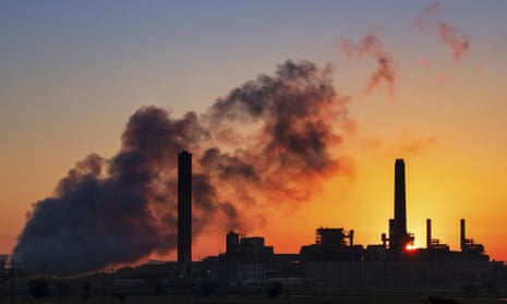 A coal-fired power plant with tall smokestacks is silhouetted against a golden yellow morning sky, as dark clouds of smoke or steam rise up from the left of the image.