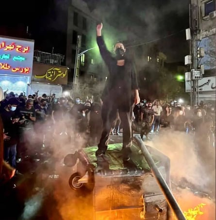 A masked woman stands on an overturned bin with her fist raised in defiance