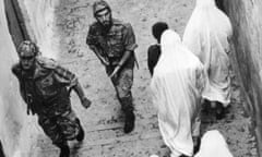 Two armed soldiers in Western combat gear pass women in traditional dress in a narrow street.