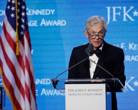 Former Fed chair Jerome Powell stands at a podium, giving a speech in front of a blue background and American flag.