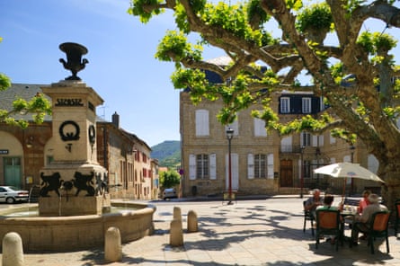 A village square with fountain and people sitting under a parasol under a tree.