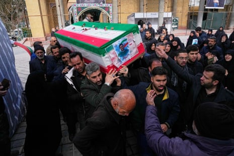 Men in black shoulder a coffin decorated in the red, white and green of the Iranian flag. On the front of the coffin is a photo of Mehdi Hossein, a civilian killed in a strike.