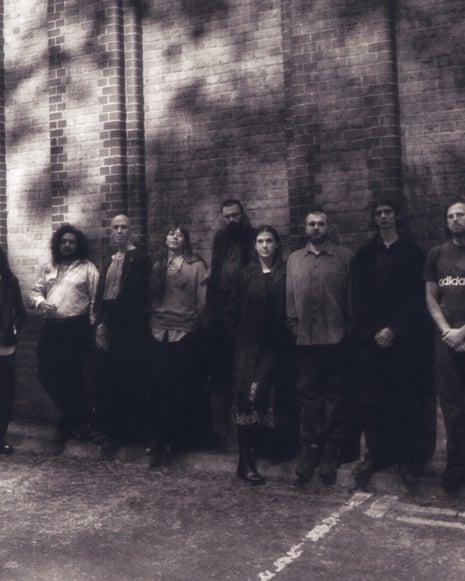 Nine members of Shovel Dance Collective lined up against a tall brick wall, dappled by shadow from a tree