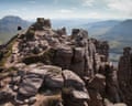 A hiker walks down a path on a rocky summit, with valley and further hills behind