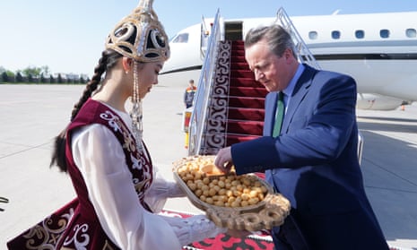A woman in central Asian heritage-style dress serves David Cameron a tray of finger food on the runway in front of red-carpeted stairs to his jet plane.