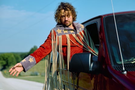 A man hanging out of the passenger seat of a car on a road in a red, white and blue tasselled jacket