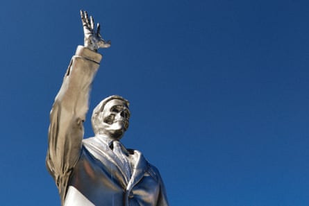 A melting silver statue of Ron DeSantis with a skull-like face raises its arm against a blue sky