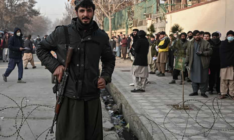 A Taliban fighter stands guard at a checkpoint as people queue to enter the passport office in Kabul