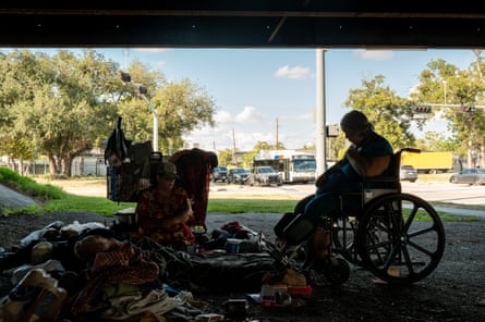 silhouette of two people, one in a wheel chair and one resting on the floor, under an overpass with their belongings