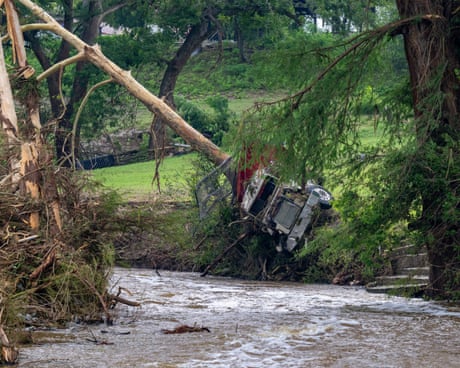 truck lays on side in grass under a tree branch next to riverbank