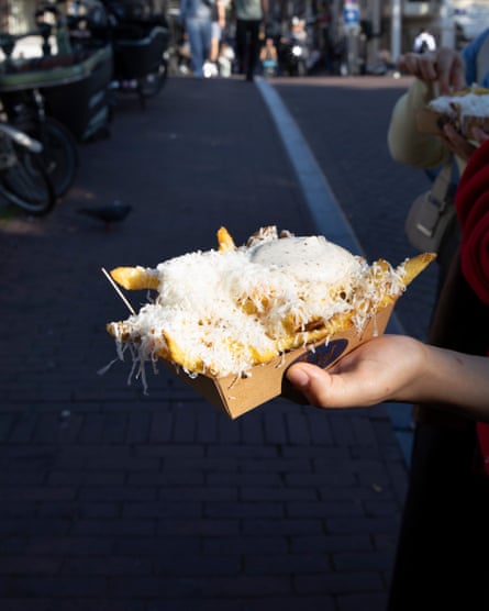 A person holding a cardboard takeaway box full of chips and covered in cheese, with a bridge in the background