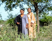 Two Wayuu women stand in an overgrown field under a tree with yellow flowers