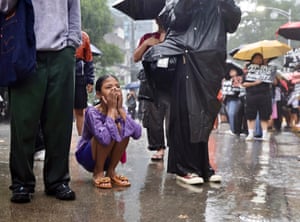 A young girl crouches on the ground in the rain, part of a group viewing a demonstration in front of a police station